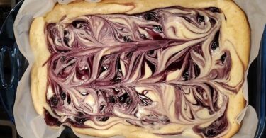 A close-up photo of a slice of Blueberry Swirl Lemon Cheesecake Bars on a white plate with a fork. The cheesecake bar has a graham cracker crust and is topped with a swirl of blueberry preserves and a sprinkle of lemon zest.