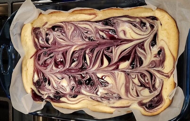 A close-up photo of a slice of Blueberry Swirl Lemon Cheesecake Bars on a white plate with a fork. The cheesecake bar has a graham cracker crust and is topped with a swirl of blueberry preserves and a sprinkle of lemon zest.