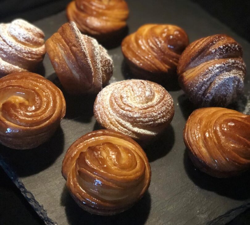 A close-up photo of a freshly baked cruffin with layers of flaky pastry and cinnamon-sugar filling, on a rustic background.